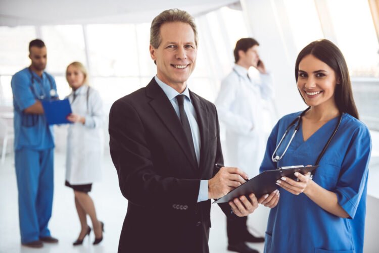 Handsome mature businessman and beautiful young doctor are looking at camera and smiling while standing in the hospital hall