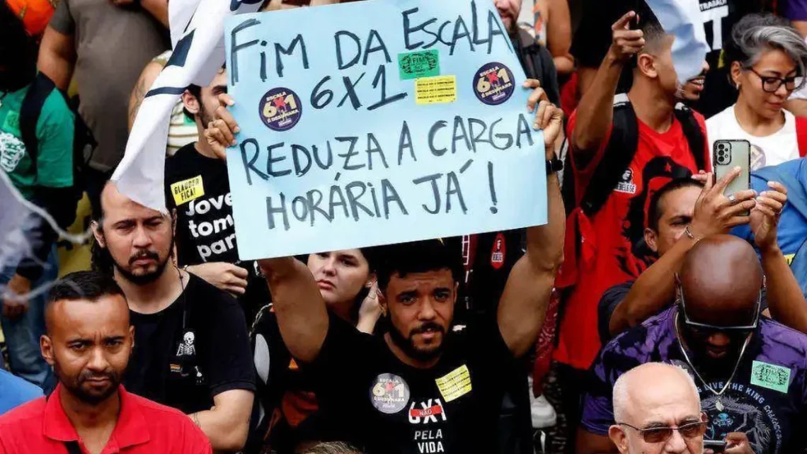 Homem no centro da imagem segura cartaz com texto "Fim da escala 6x1, reduza a carga horária já!" em meio a grupo de manifestantes. Pessoas ao redor seguram bandeiras e celulares, algumas usam camisetas pretas com mensagens relacionadas ao trabalho.
Manifestação pelo fim da escala 6x1 em 2024, na Cinelândia, Rio de Janeiro (RJ) - Tânia Rêgo/Agência Brasil